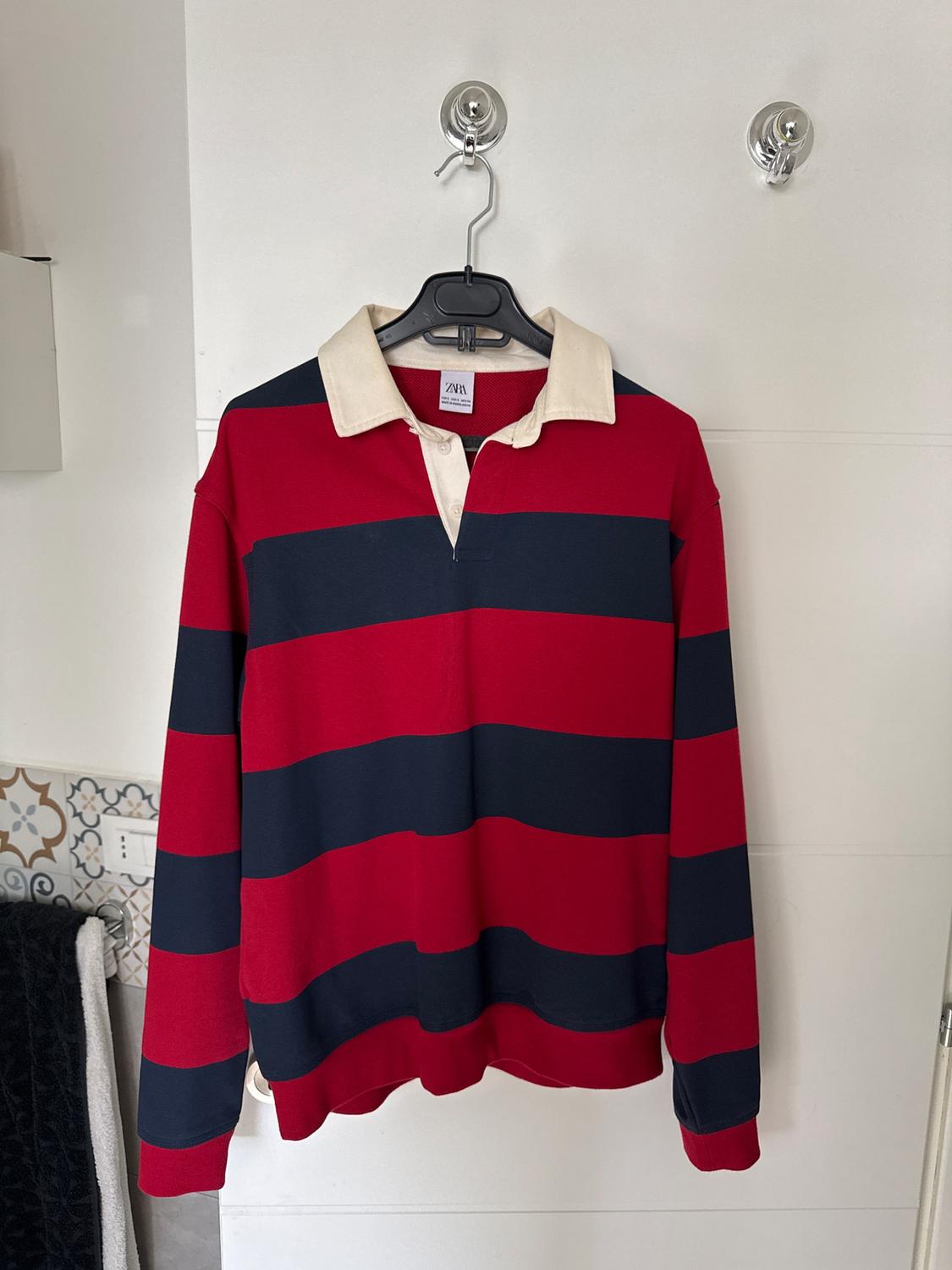 Red and navy blue horizontal striped shirt with a white collar, hanging on a black hanger against a white wall.