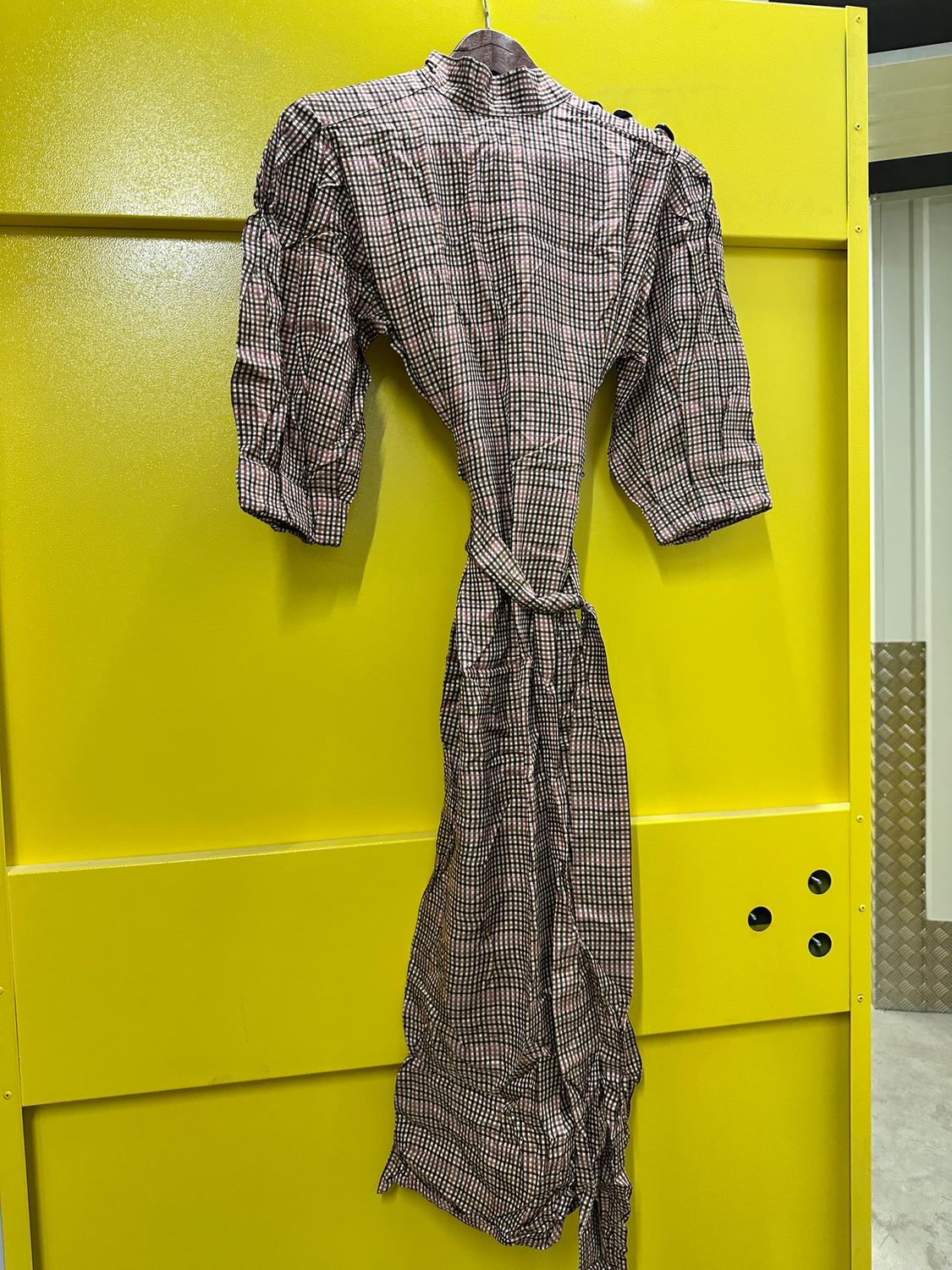 Checked dress hanging on a yellow door. Puffed sleeves and a high neck, with a bow at the waist. Industrial setting.