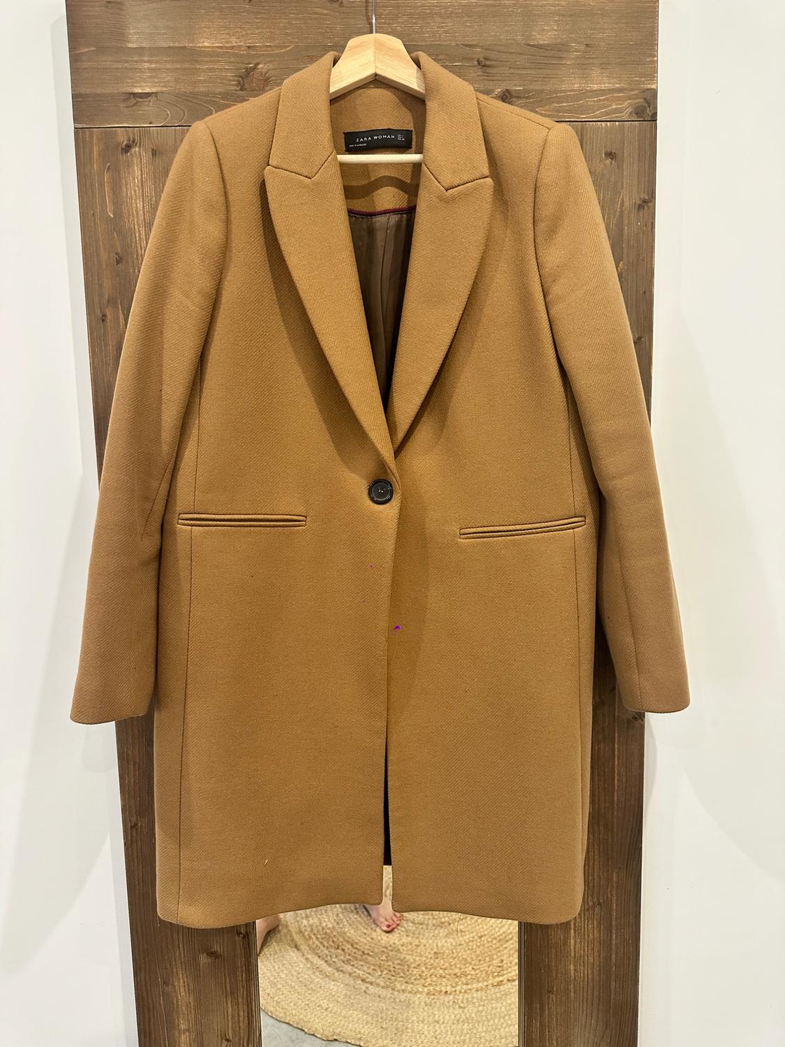 Camel-coloured coat hanging on a hanger against a wooden panel. ZARA WOMAN label. The reflection below shows bare feet and a rug in a casual setting.