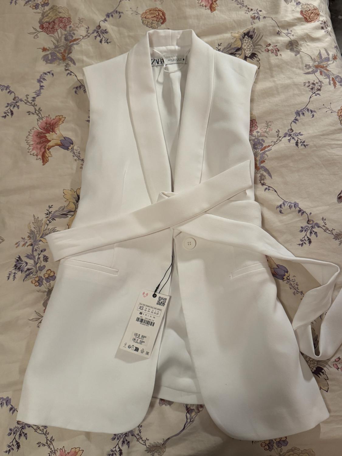 White sleeveless vest with a belt over floral fabric. Visible tag with price and size XS.