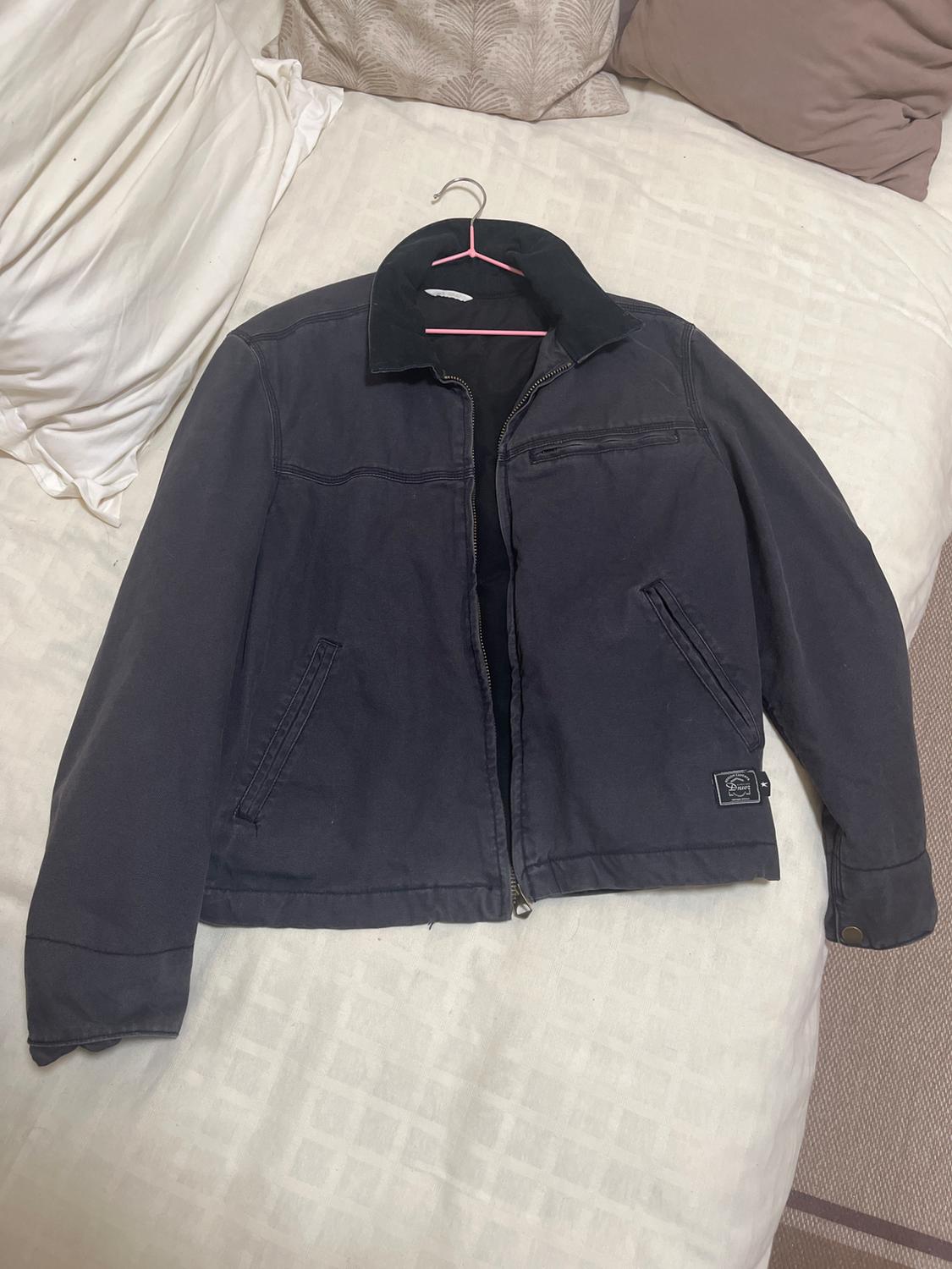 Black zip-up, high-neck jacket, hanging on a pink hanger over a bed with white sheets and beige cushions.