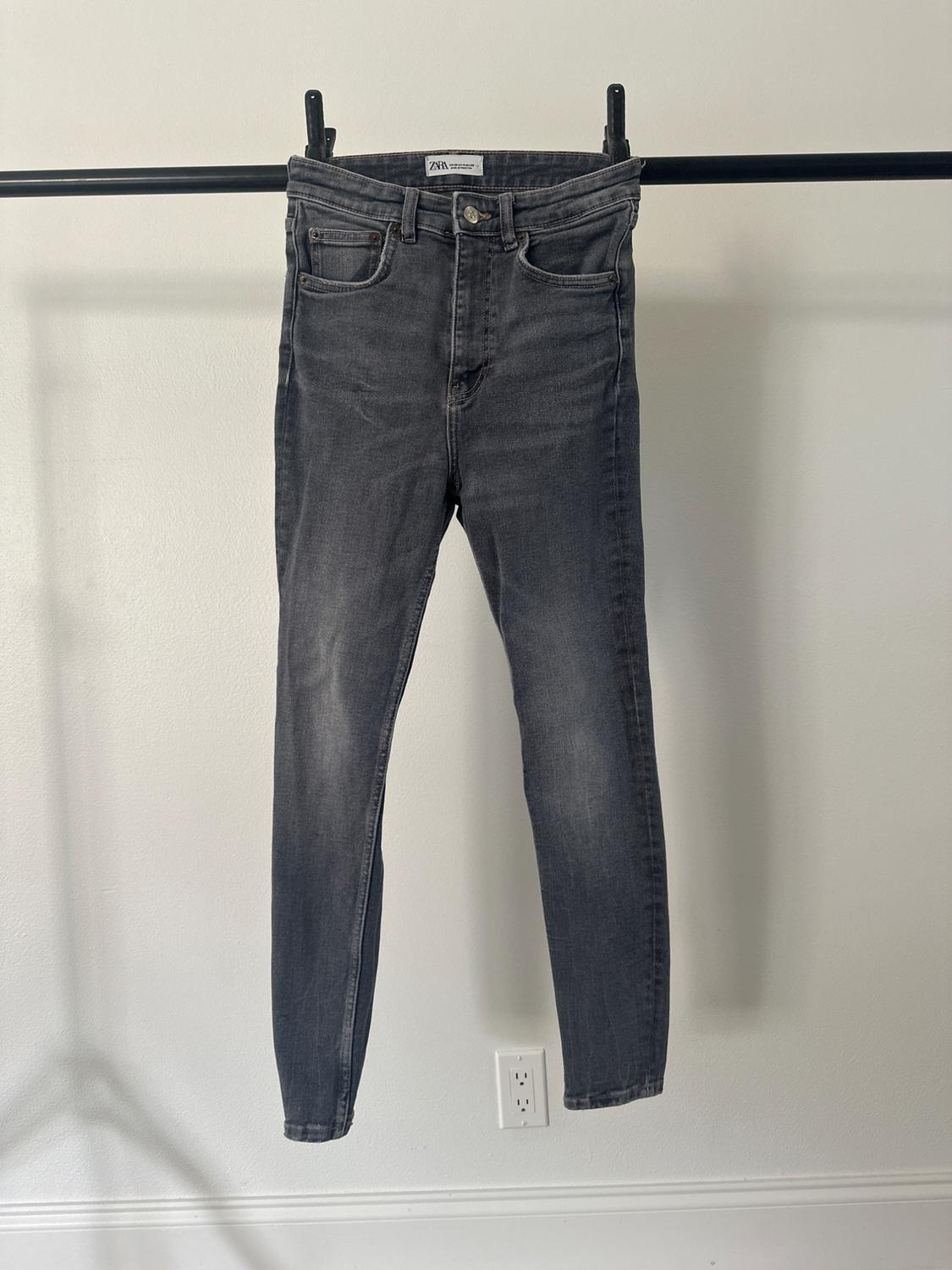 Grey jeans hanging on a black rail against a white wall. Label visible on the waistband with the text "Zara".