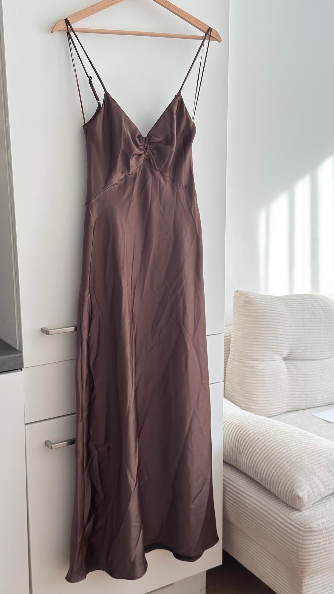 Brown spaghetti strap dress hanging on a wooden hanger, above a white wardrobe next to a light-coloured sofa.