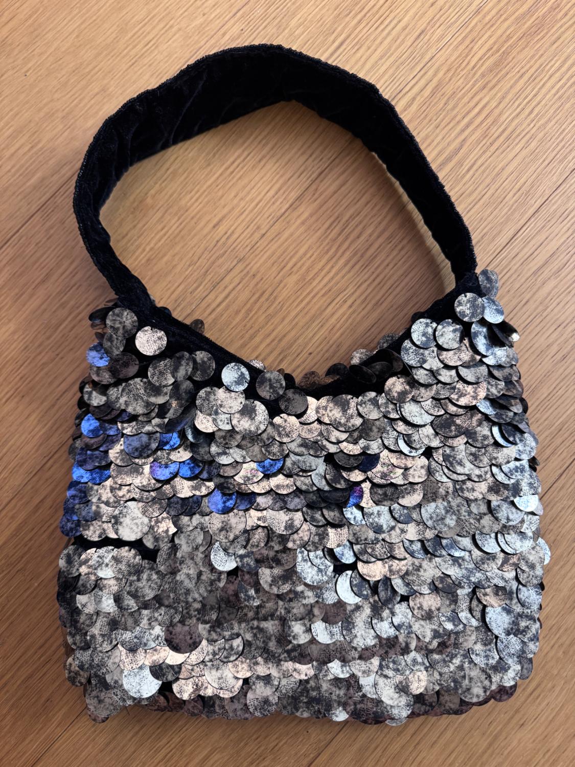 Silver and blue sequined handbag on a black background, placed on a light wooden surface.