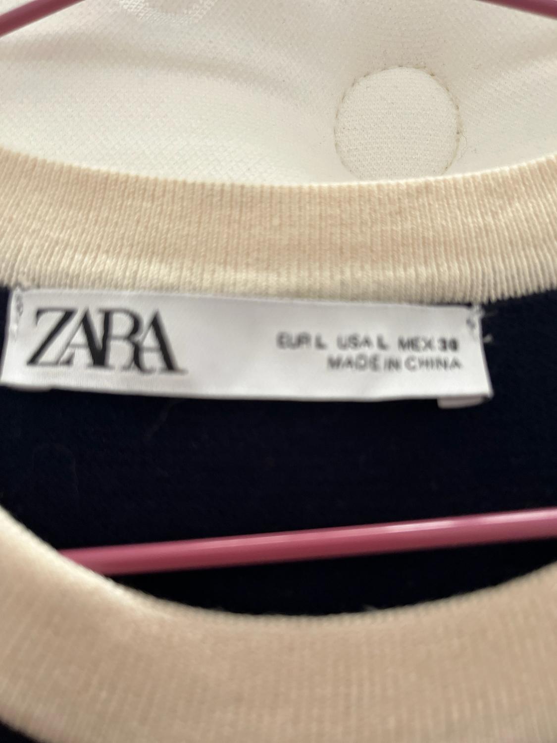 Ετικέτα με το κείμενο ZARA EUR L USA L MEX 30 MADE IN CHINA σε ένα σκουρόχρωμο
ένδυμα με μπεζ γιακά, κρεμασμένο σε μια ροζ κρεμάστρα.