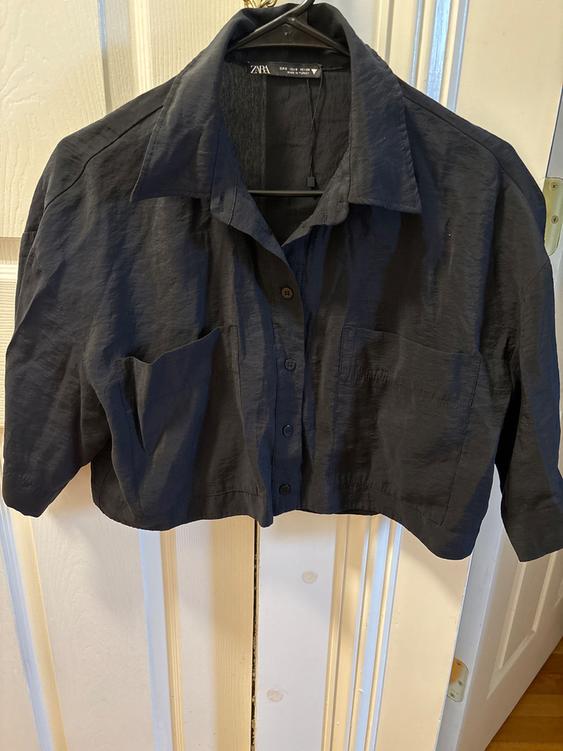 Black short-sleeved shirt hanging on a hanger, with front buttons and a chest pocket. Visible Zara tag.