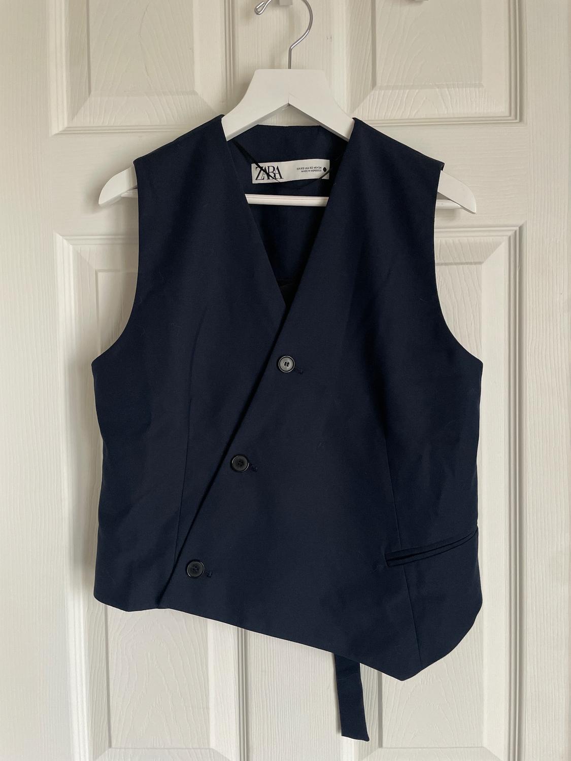 Black sleeveless waistcoat with a crossover design and three buttons, hanging on a white hanger in front of a white door. Label visible.