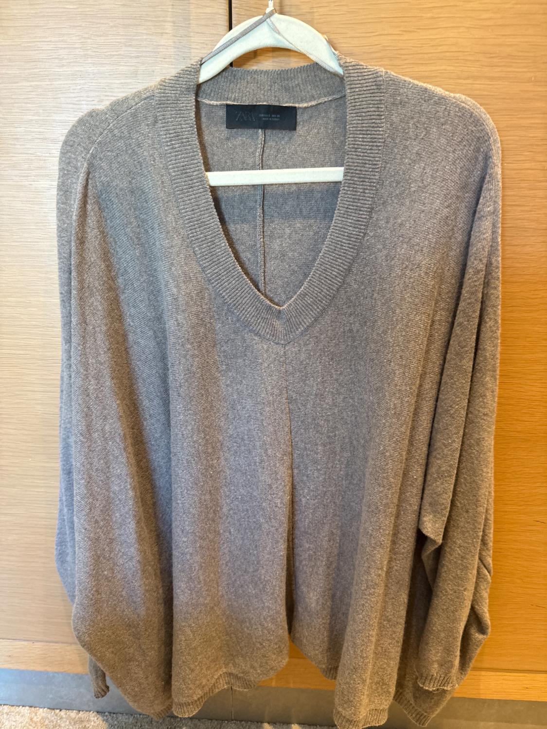 Grey knitted V-neck jumper hanging on a wooden hanger. Light wooden background. Visible label on the collar.