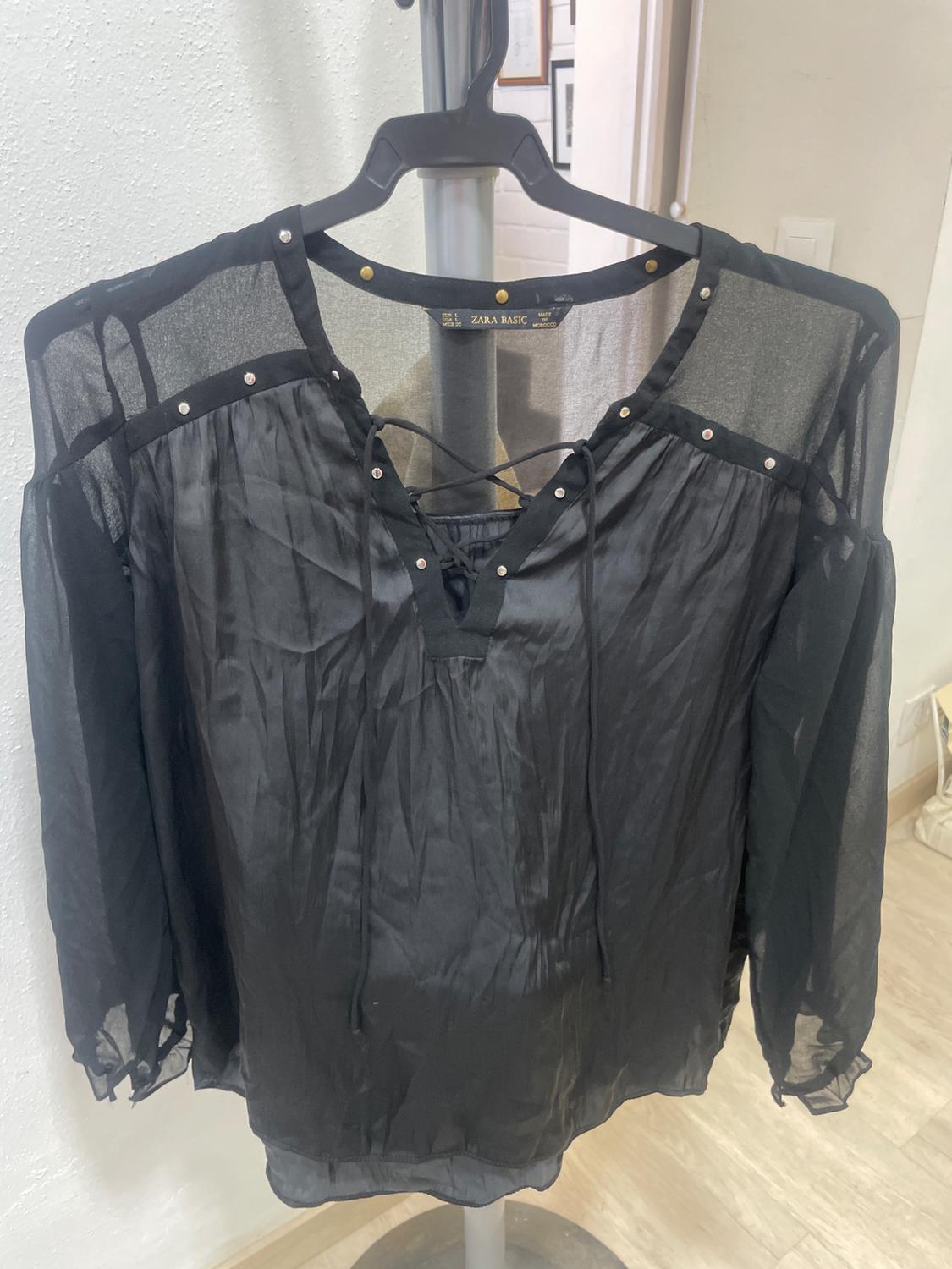 Semi-sheer black blouse with stud detailing and lace-up neckline, hanging on a hanger inside a room.