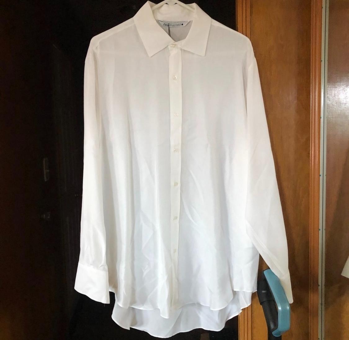 White long-sleeved shirt hanging on a hanger, with a wooden background and a mirror reflecting part of the garment.
