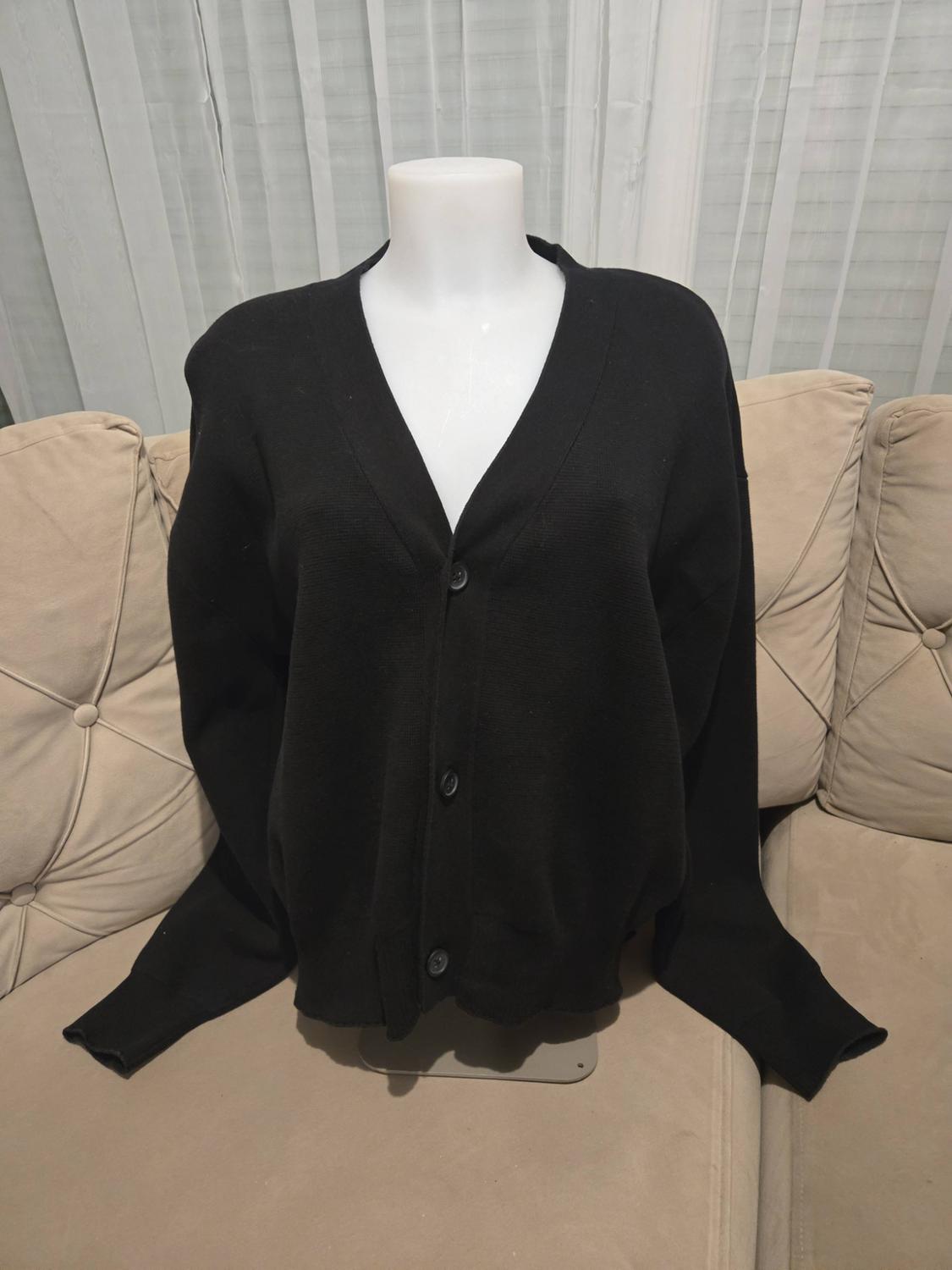 Black knitted button-up V-neck cardigan on a white mannequin, on a beige sofa with curtains in the background.
