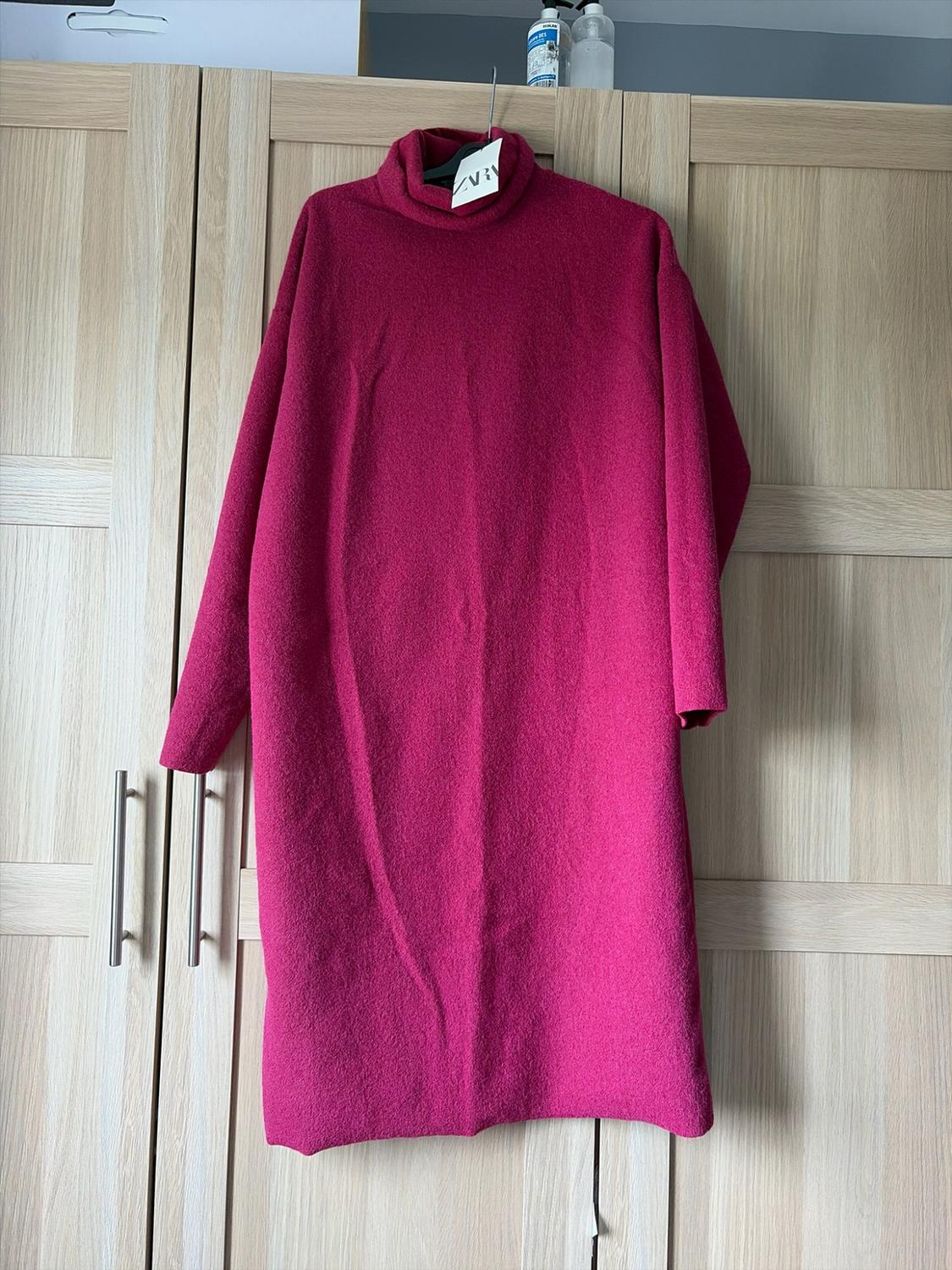 Fuchsia pink long-sleeved, high-neck dress hanging in a light wood wardrobe. Relaxed atmosphere. Label with ZARA text.