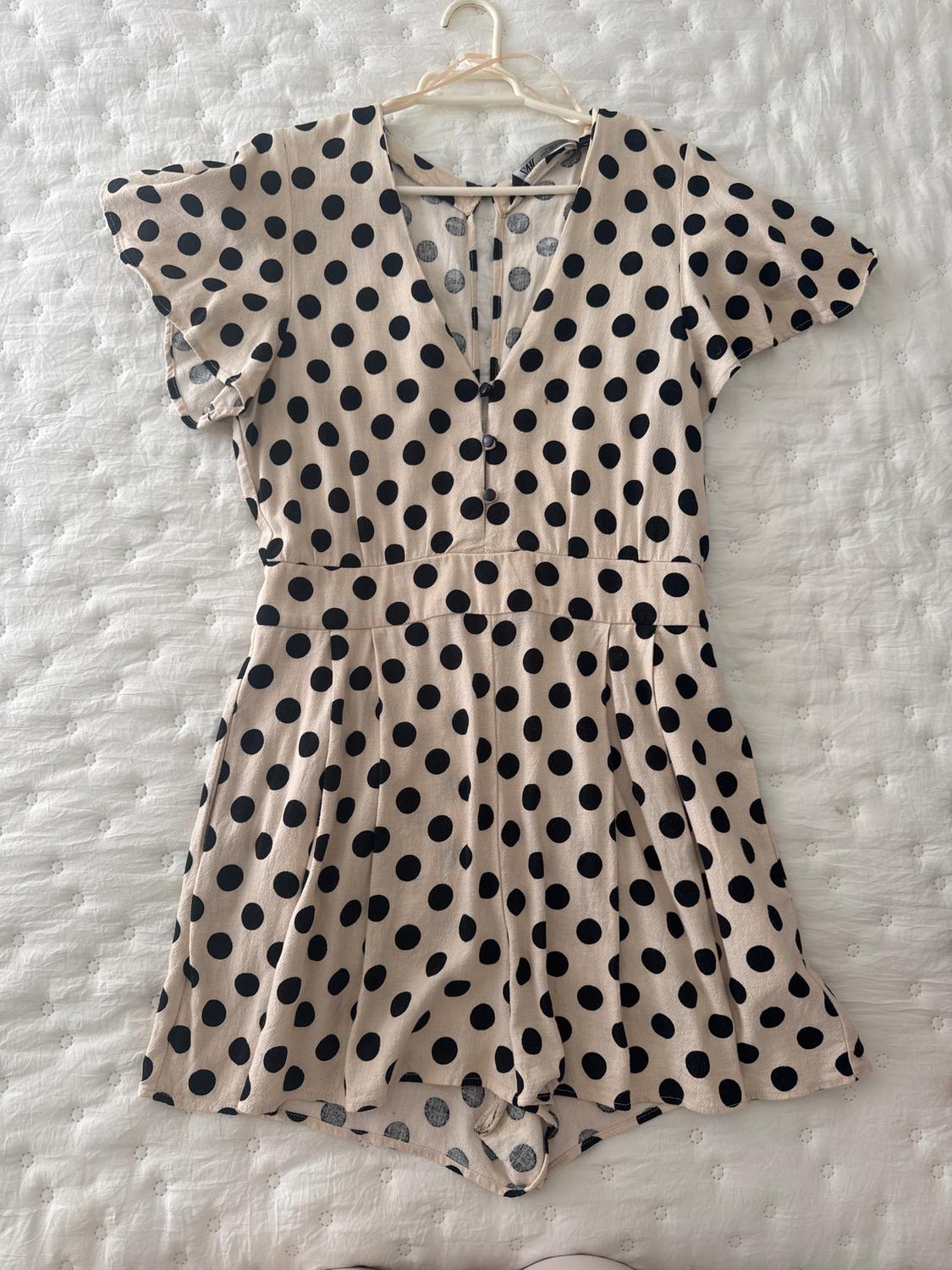Beige jumpsuit with black polka dots, short sleeves and V-neck, hanging on a hanger over a white quilted bedspread.