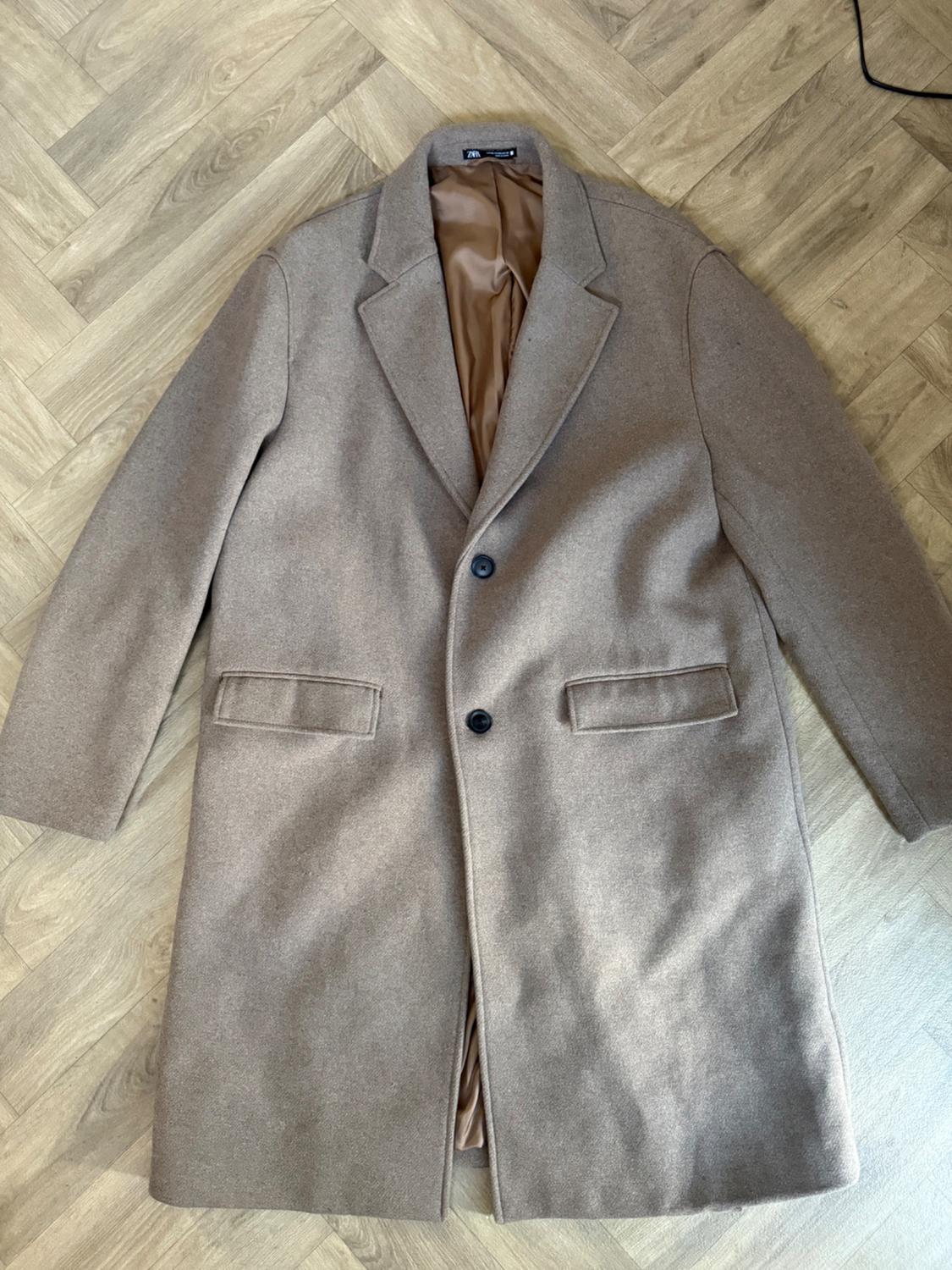 Long beige coat with a lapel collar, two dark buttons, and flap pockets, laid out on a herringbone wooden floor.