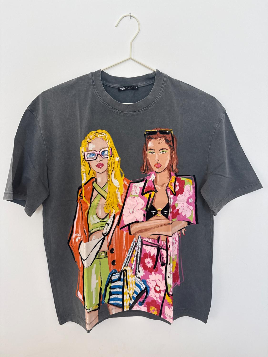 Σκούρο γκρι T-shirt κρεμασμένο σε κρεμάστρα. Ξεχωρίζει ένα πολύχρωμο τύπωμα δύο γυναικών με γυαλιά ηλίου και καλοκαιρινά ρούχα.