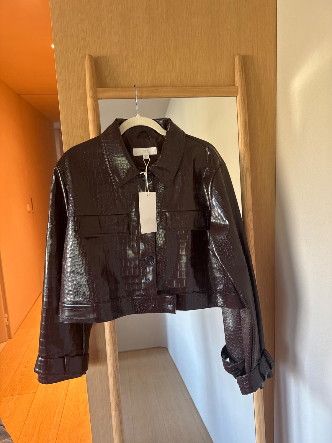 Brown cropped jacket with a crocodile texture hanging on a wooden mirror. White label visible on the collar.