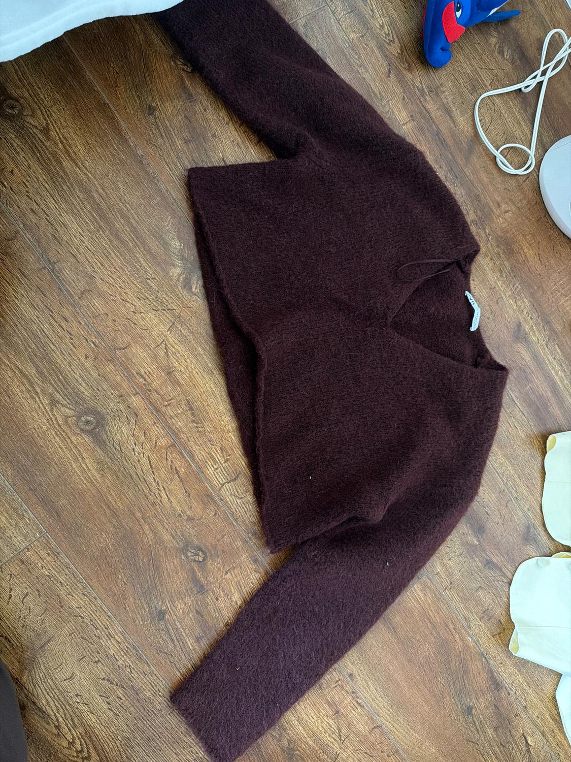 Dark brown wool V-neck jumper on a wooden floor. In the background, a blue toy and white cables.