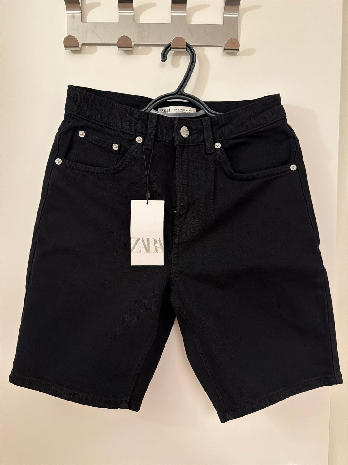 Black denim shorts hanging on a hanger on a metal wall rack. White label with the text ZARA.