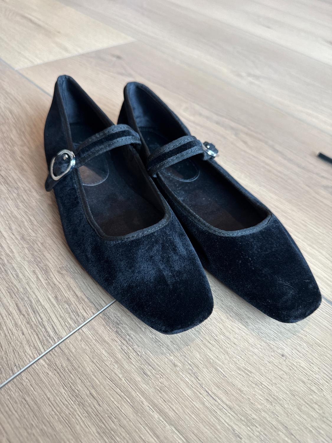 Pair of black velvet Mary Jane flat shoes with a strap and silver buckle, on a light wood floor.