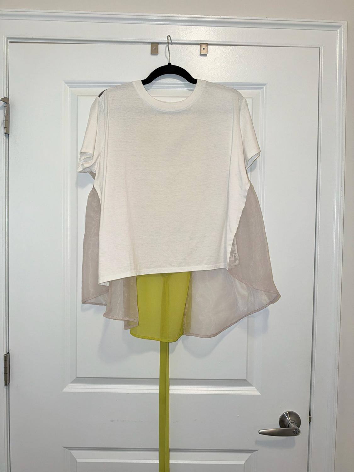 White short-sleeve T-shirt with a semi-transparent back panel and yellow detail, hanging on a hanger over a white door.