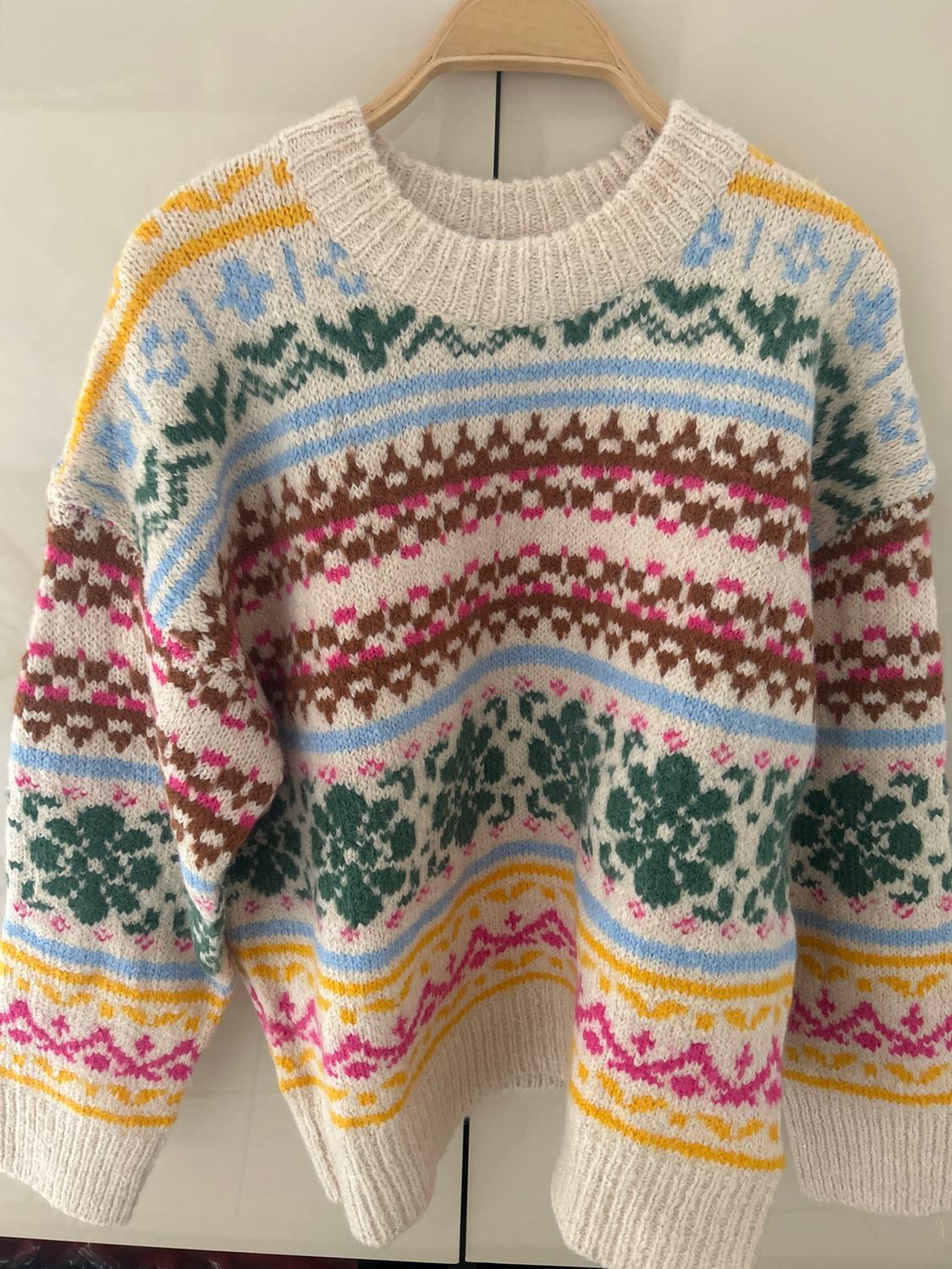 Knitted jumper hanging on a hanger, with a colourful floral and geometric pattern design in shades of green, blue, pink, brown, and yellow.