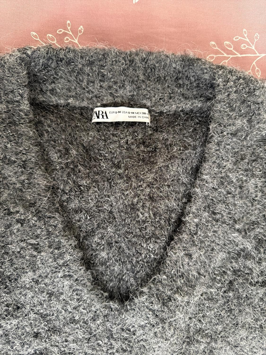 Detail of a grey knit V-neck sweater on a pink background. Text: ZARA EUR S-M USA S-M MEX 38-4 MADE IN CHINA.