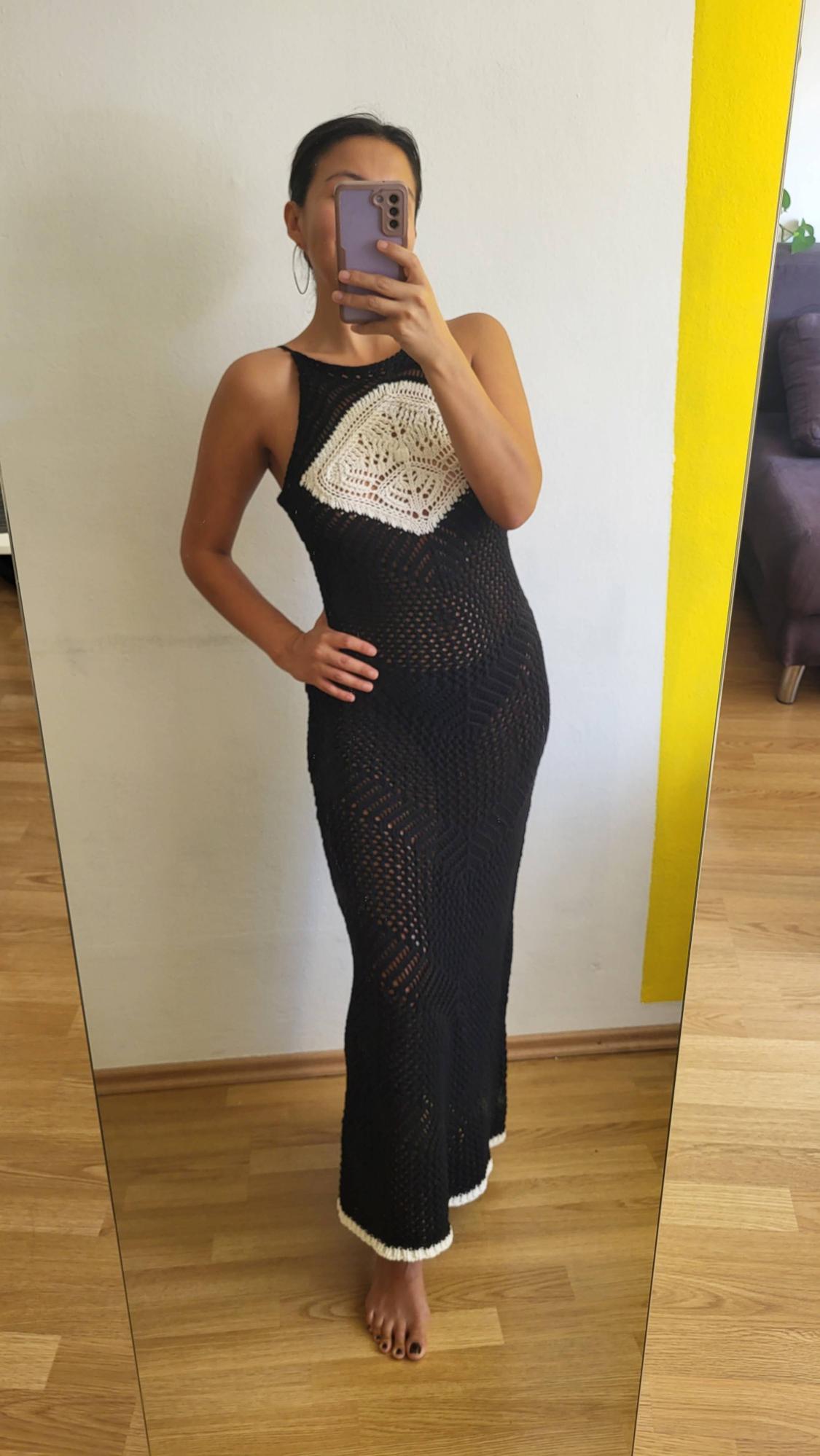 Woman in front of a mirror, wearing a long black crochet dress with white detail on the chest. She is holding a mobile phone.