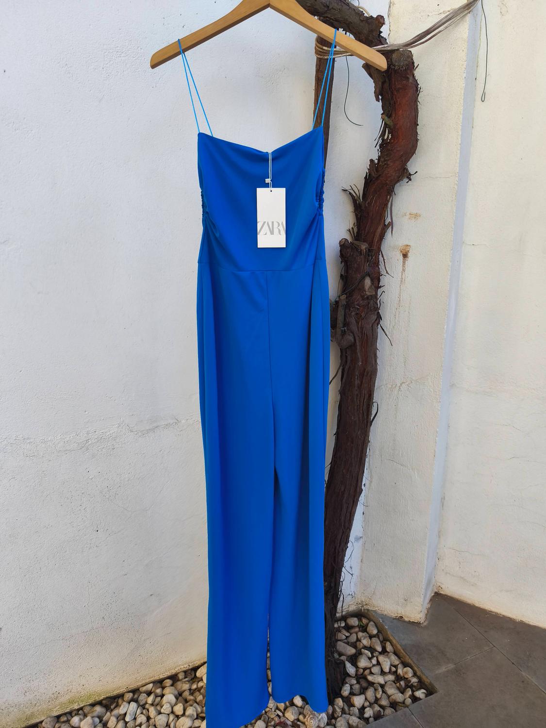 Blue dungarees with thin straps, hanging on a wooden hanger. Visible label with text "ZARA". White wall and stone background.