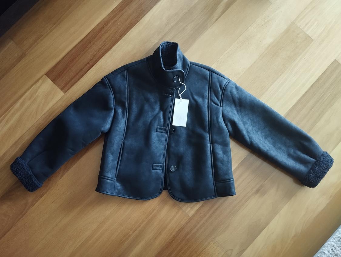 Black synthetic jacket with wool lining, white hanging tag, on a light wooden floor.