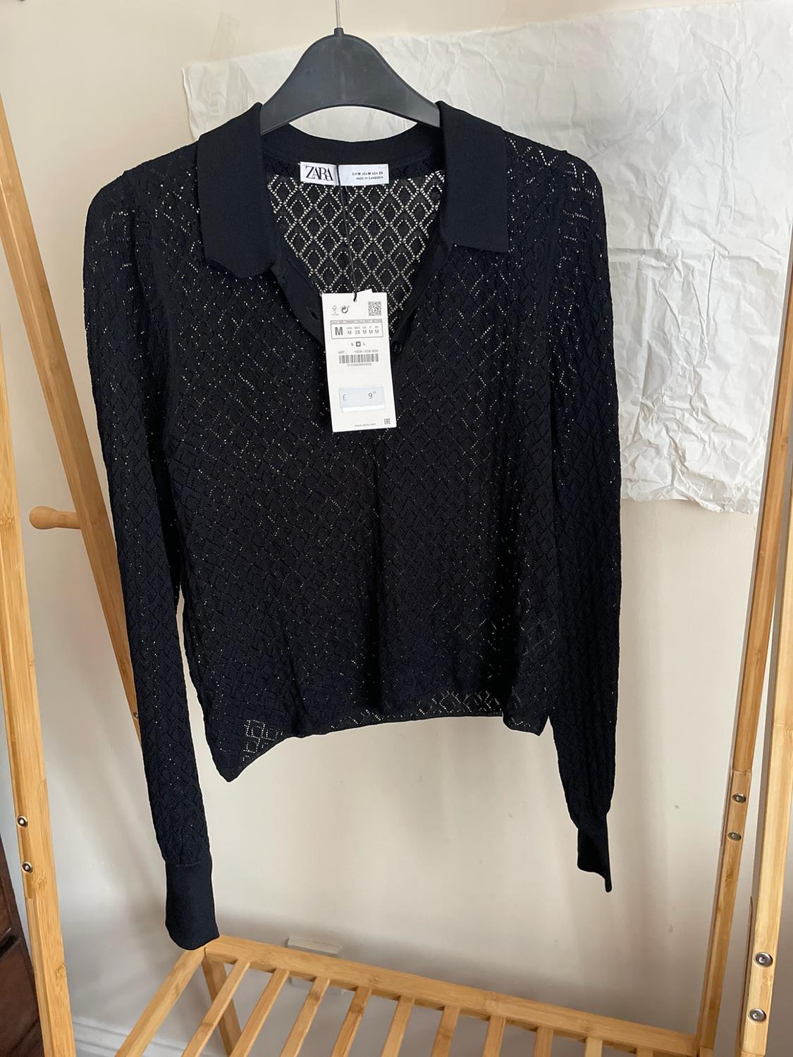 Black openwork polo shirt hanging on a wooden clothes rail. Label with text: ZARA EUR M USA M MEX 28 MADE IN CAMBODIA M.
