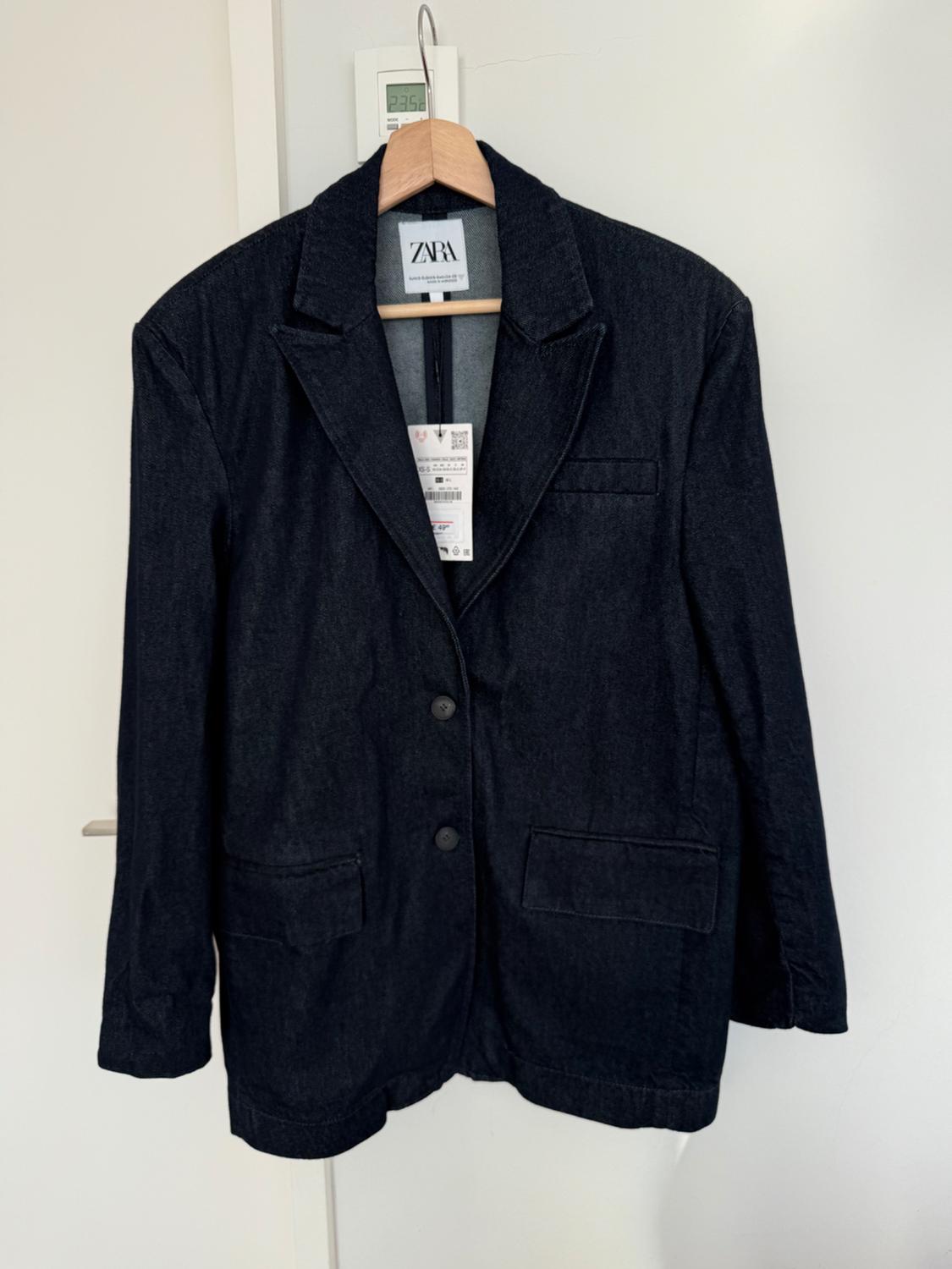 Black denim jacket hanging on a wooden hanger. Visible label with the text "ZARA". Minimalist white background.