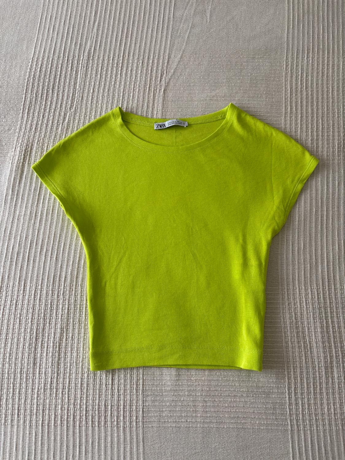 Κοντό t-shirt σε λαχανί χρώμα πάνω σε μια μπεζ ανάγλυφη επιφάνεια. Ορατή ετικέτα στο λαιμό με την επωνυμία "ZARA".