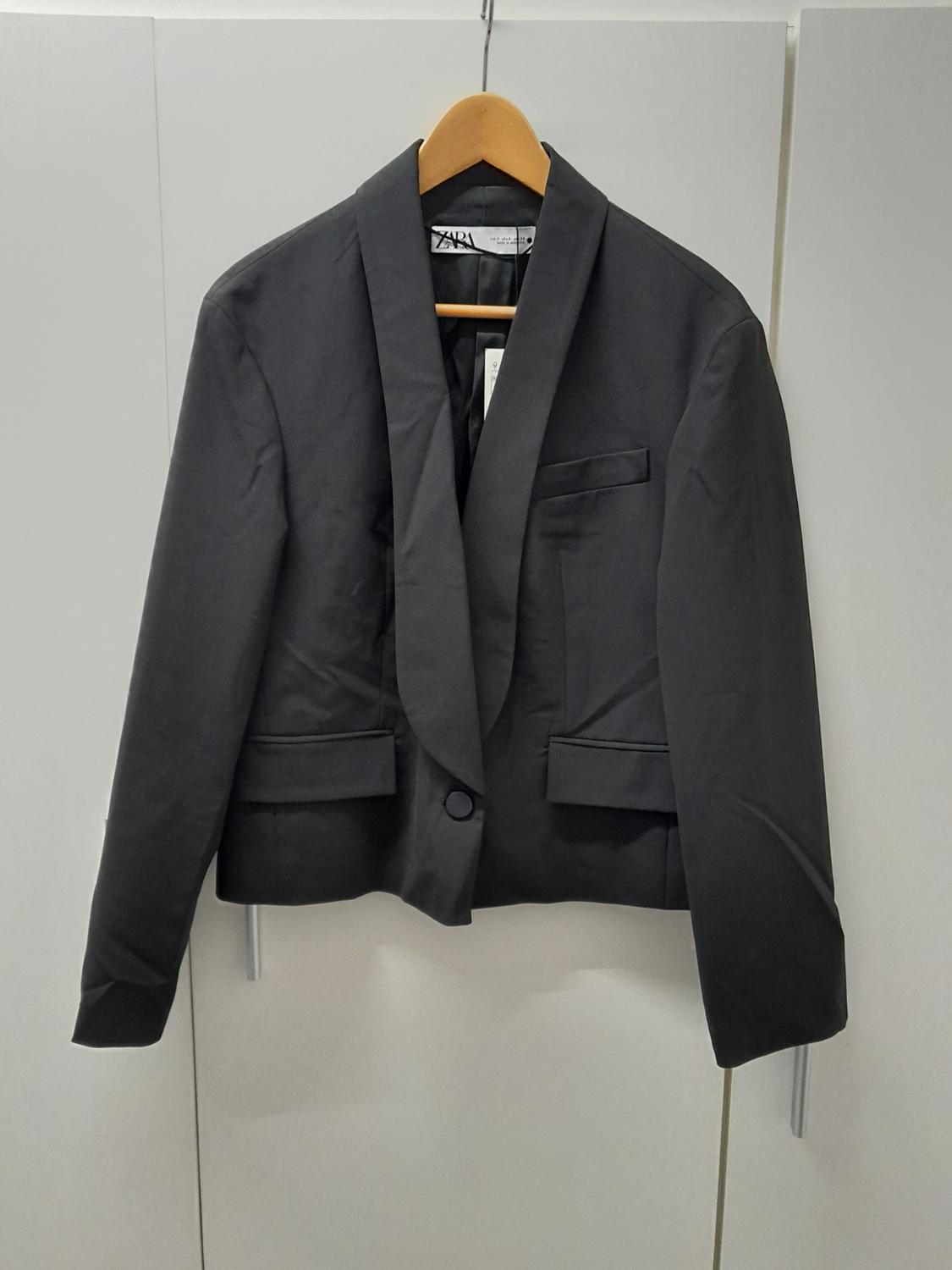 Black suit jacket hanging on a wooden hanger, with a wide lapel and one button, against a white wardrobe background.