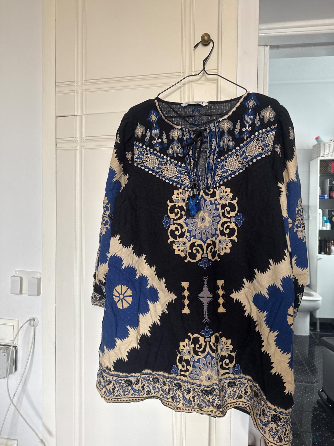 Blouse hanging on a hanger, with a blue and beige pattern on a black background, floral and geometric details, on a white door.