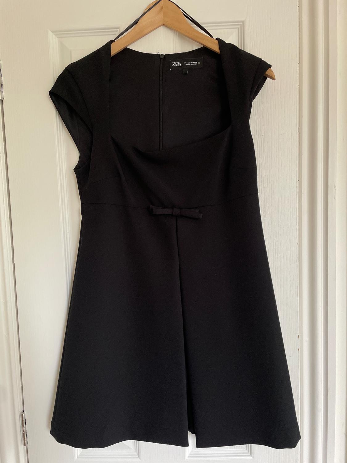 Sleeveless black dress hanging on a wooden hanger over a white door. It has a small bow at the waist.