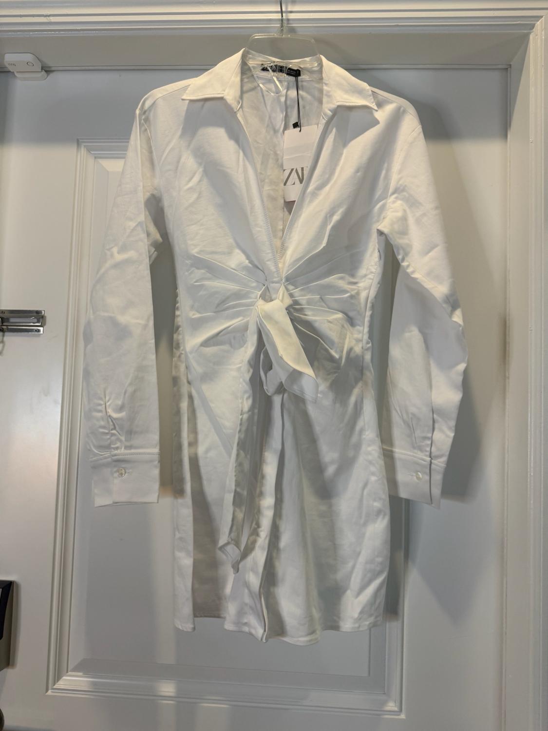 White long-sleeved shirt dress with a waist tie, hanging on a white door. Label with the text ZN.