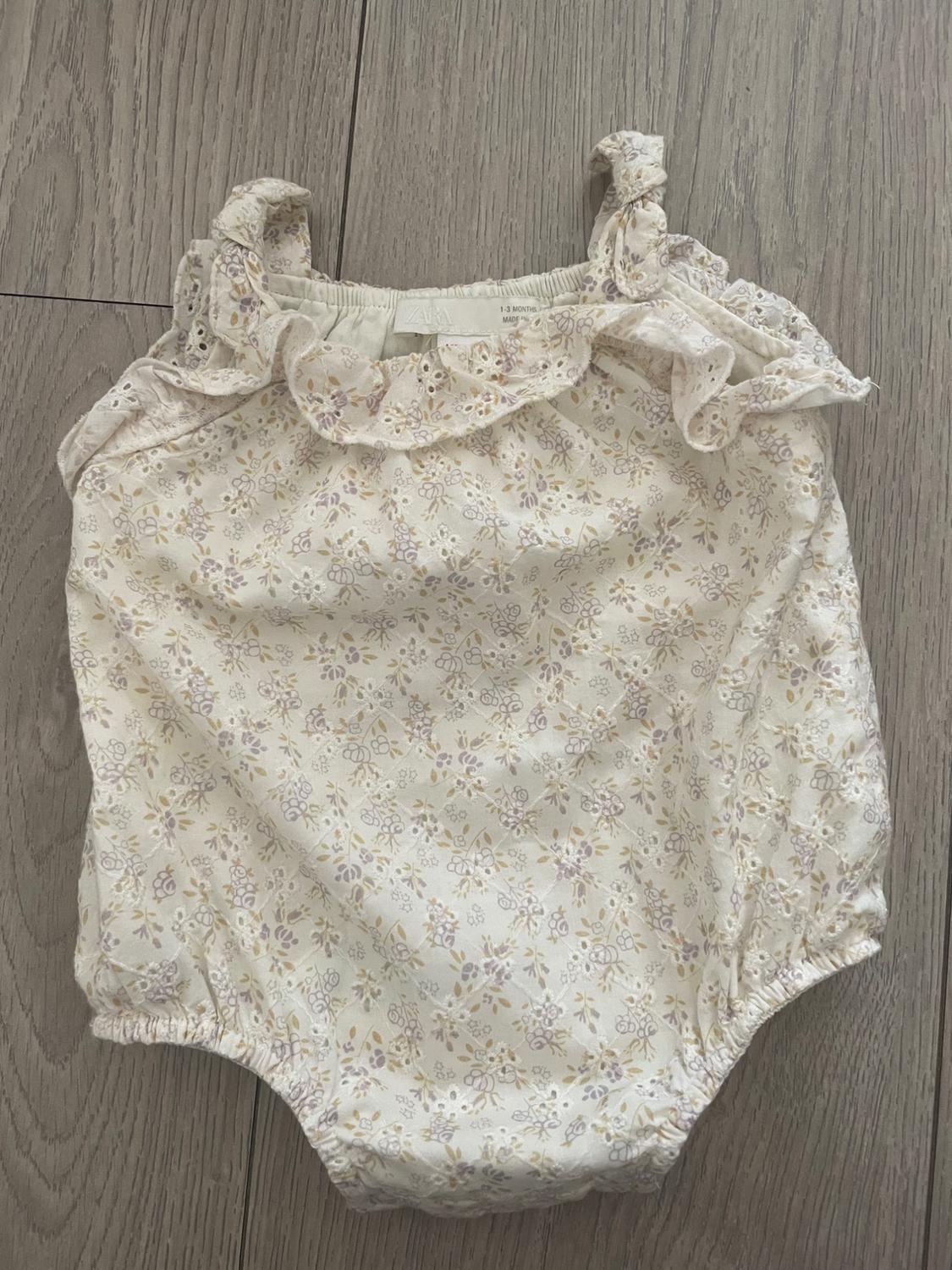 Baby bodysuit with pastel floral print, frills on the straps and neckline, on a light wood floor.