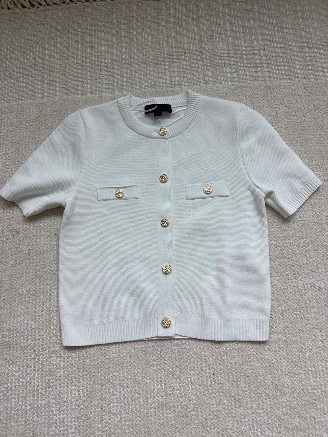 White short-sleeved jacket with gold buttons and two front pockets, on a beige textile surface.