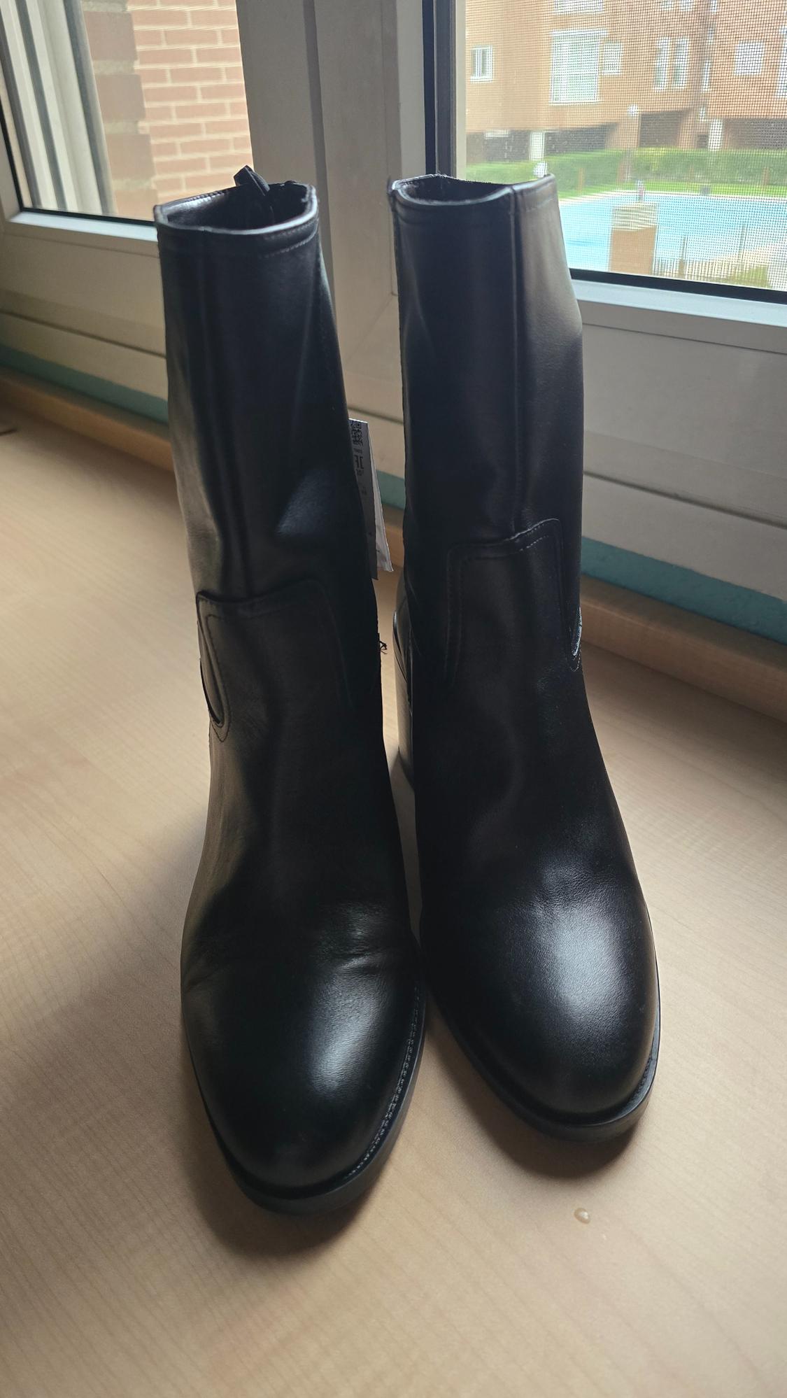 Black leather boots on a light wooden table, next to a window with a view of an outdoor courtyard.