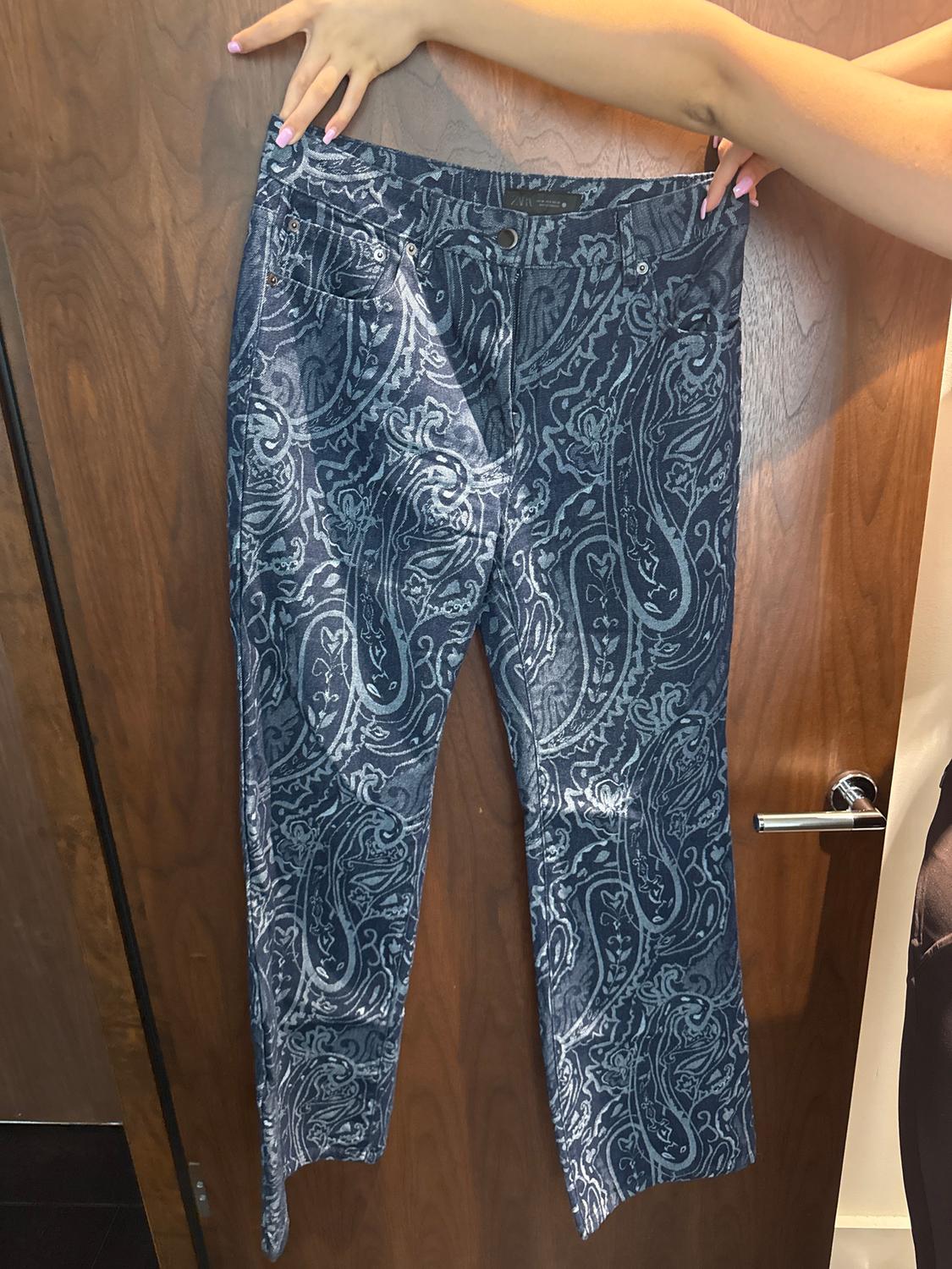 A pair of blue and white paisley-patterned denim trousers, held up by a hand with pink painted nails, with a wooden door as a background.