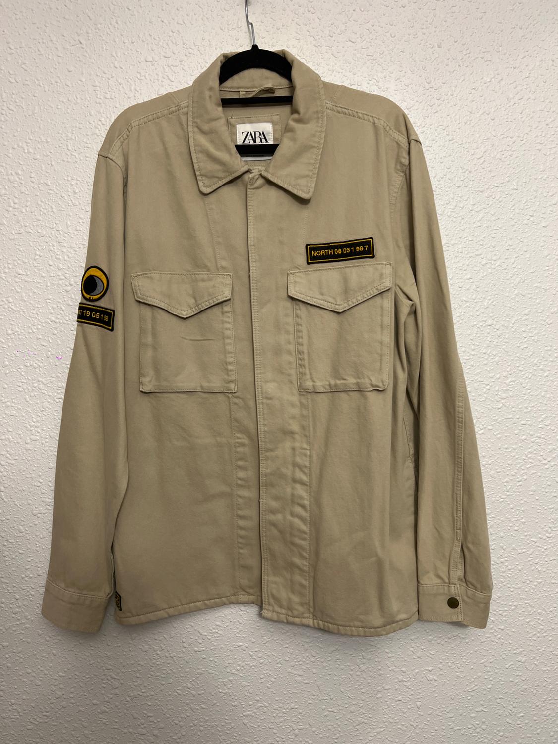 Beige jacket hanging on a hanger, with patches that say "NORTH 06 051 987" and an eye. ZARA label visible on the collar.