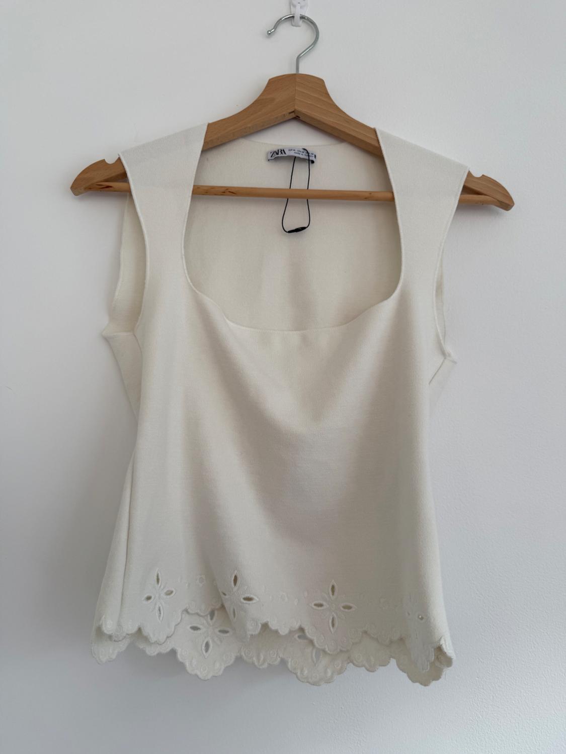 A white, sleeveless blouse with a square neckline and floral embroidery on the hem, hanging on a wooden hanger against a white background.