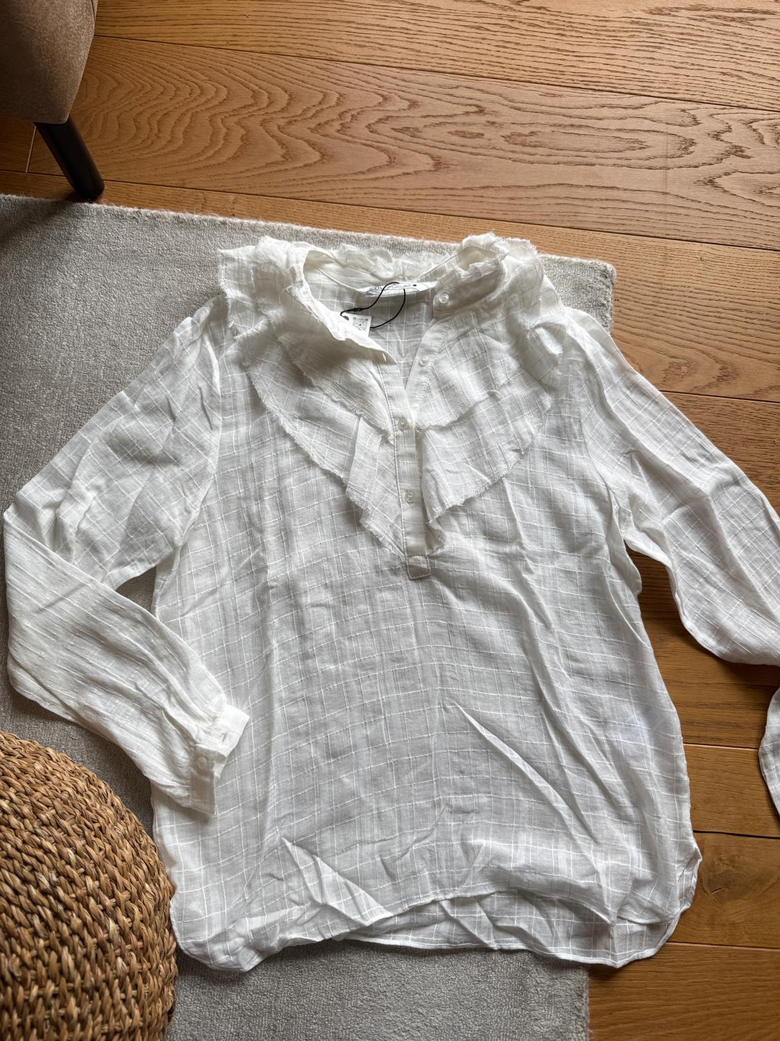 Long-sleeved white blouse with a ruffled collar, on a grey rug and light wooden floor.
