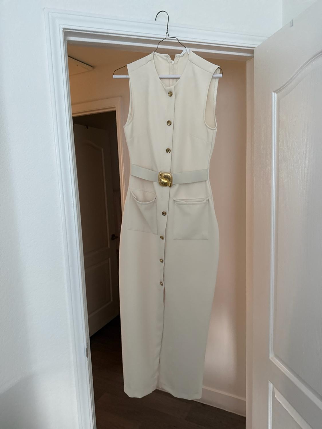 Cream-colored sleeveless midi dress with gold buttons, a belt, and pockets, hanging on a hanger in a white door frame.