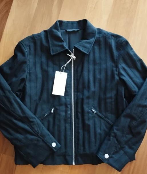 Black jacket with front zip, zip pockets, and white hanging tag, on a light wooden background.