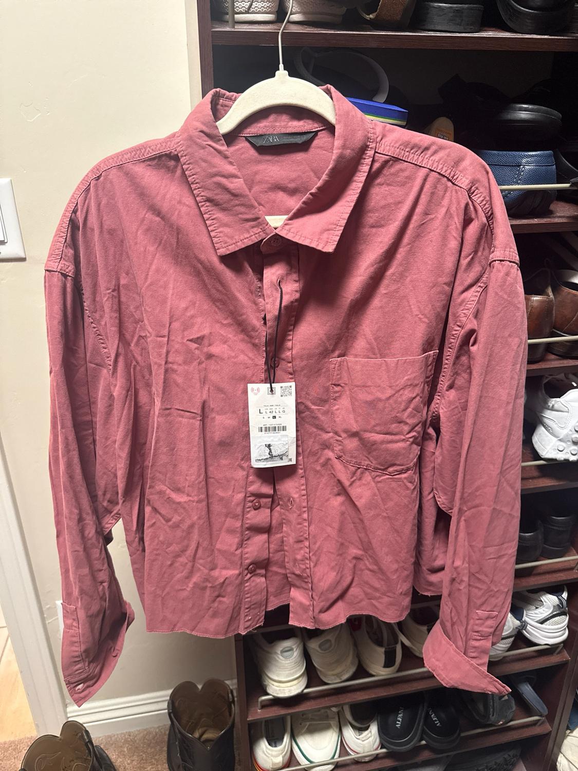 Long-sleeve pink shirt with a pocket and ZARA label, hanging on a hanger in front of a wooden shoe rack with shoes.