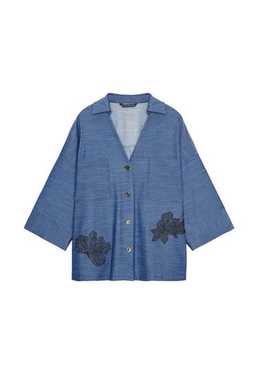 Front view of a blue shirt with a V-neck, three-quarter sleeves, patch pockets, and black floral embroidery. Metal buttons.