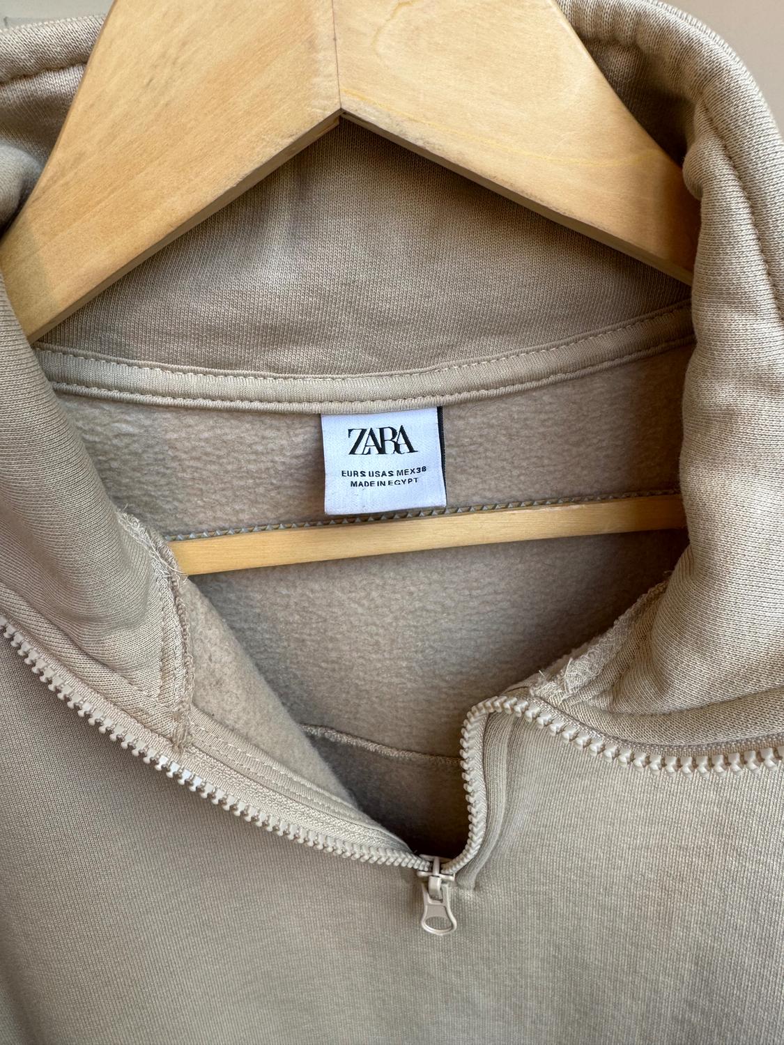 Beige zip-up sweatshirt hanging on a wooden hanger. Visible label: "ZARA EUR S USA S MEX 30 MADE IN EGYPT".