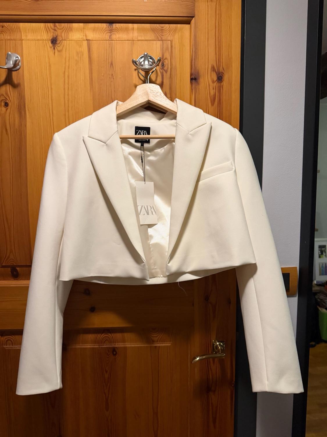 Short white blazer hanging on a wooden hanger over a door. Visible label bearing the text ZARA.
