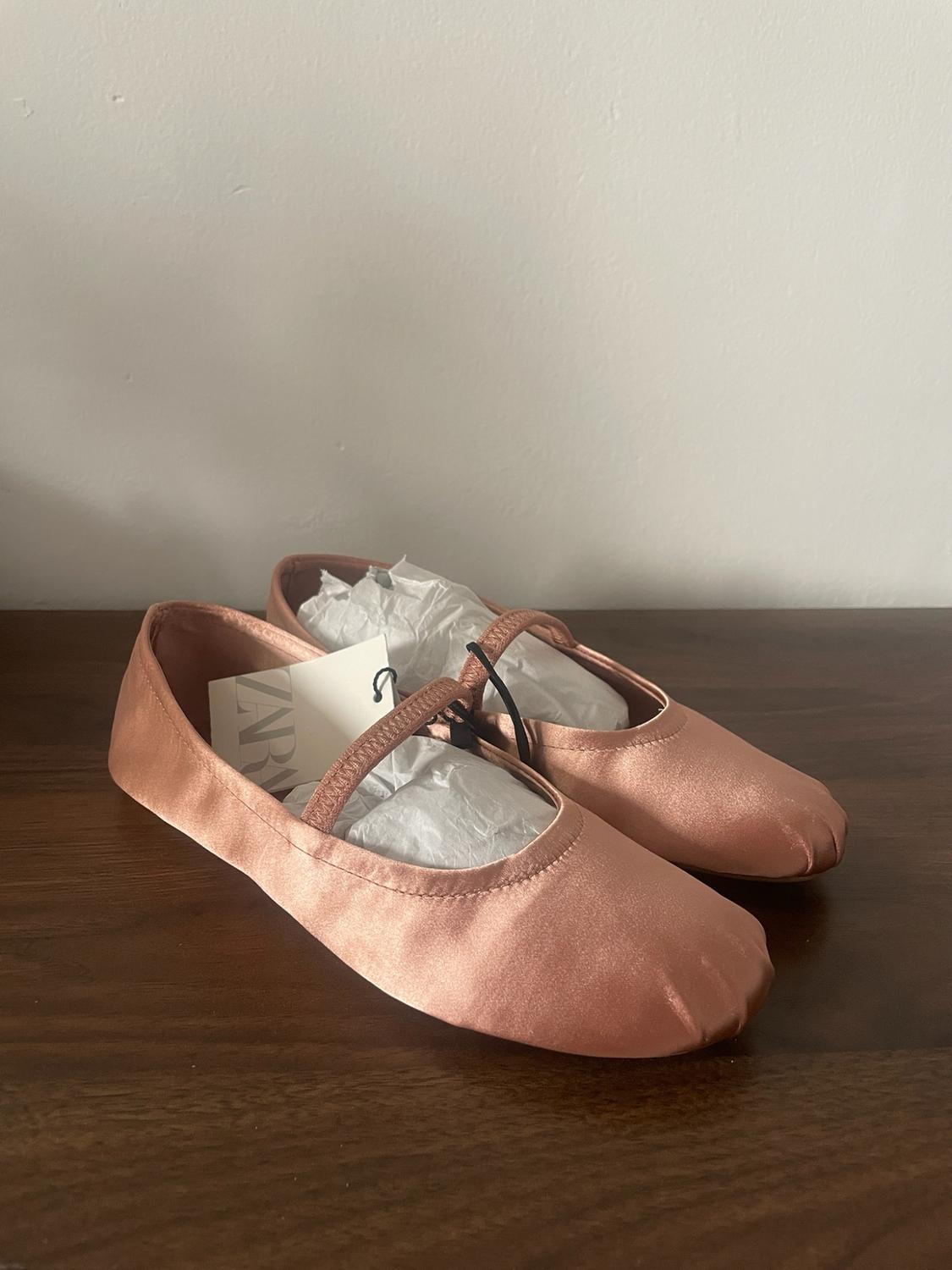 Pink satin ballet flats with an elastic strap on a wooden table. They have stuffing inside and a label with the text ZARA.